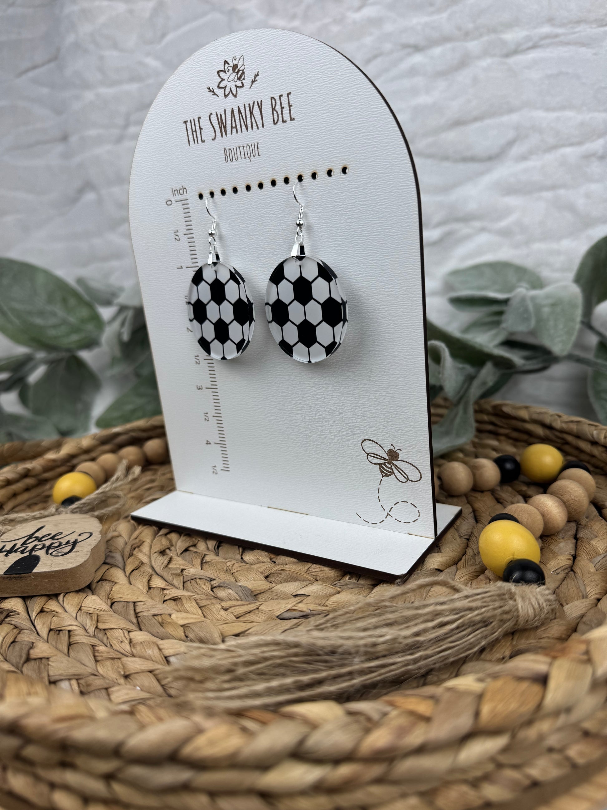 Black and white soccer ball earrings displayed on a branded card with a ruler guide, surrounded by decorative beads and greenery.