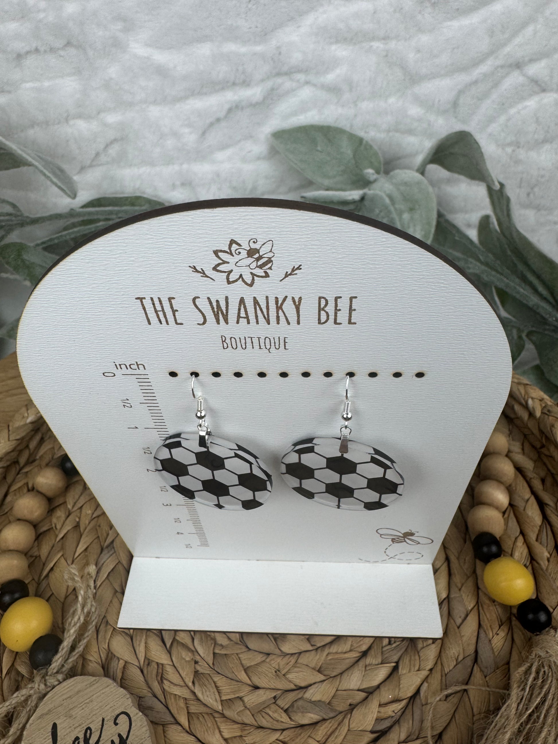 Soccer dangle earrings with black and white patterns, hanging on a branded card from The Swanky Bee Boutique, displayed on a woven basket.