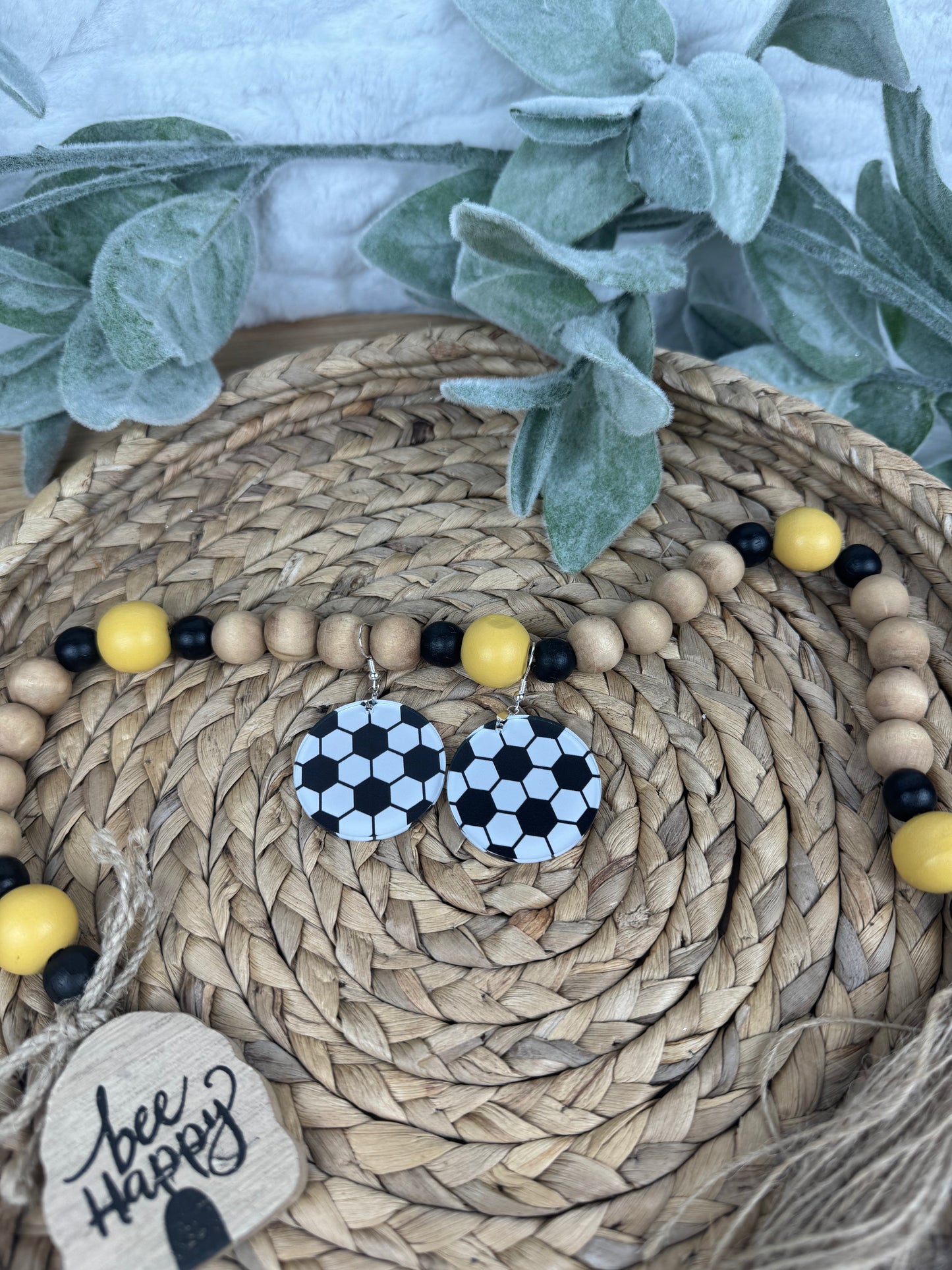 Black and white soccer ball earrings displayed on a woven basket with greenery and decorative beads, perfect for sports lovers.