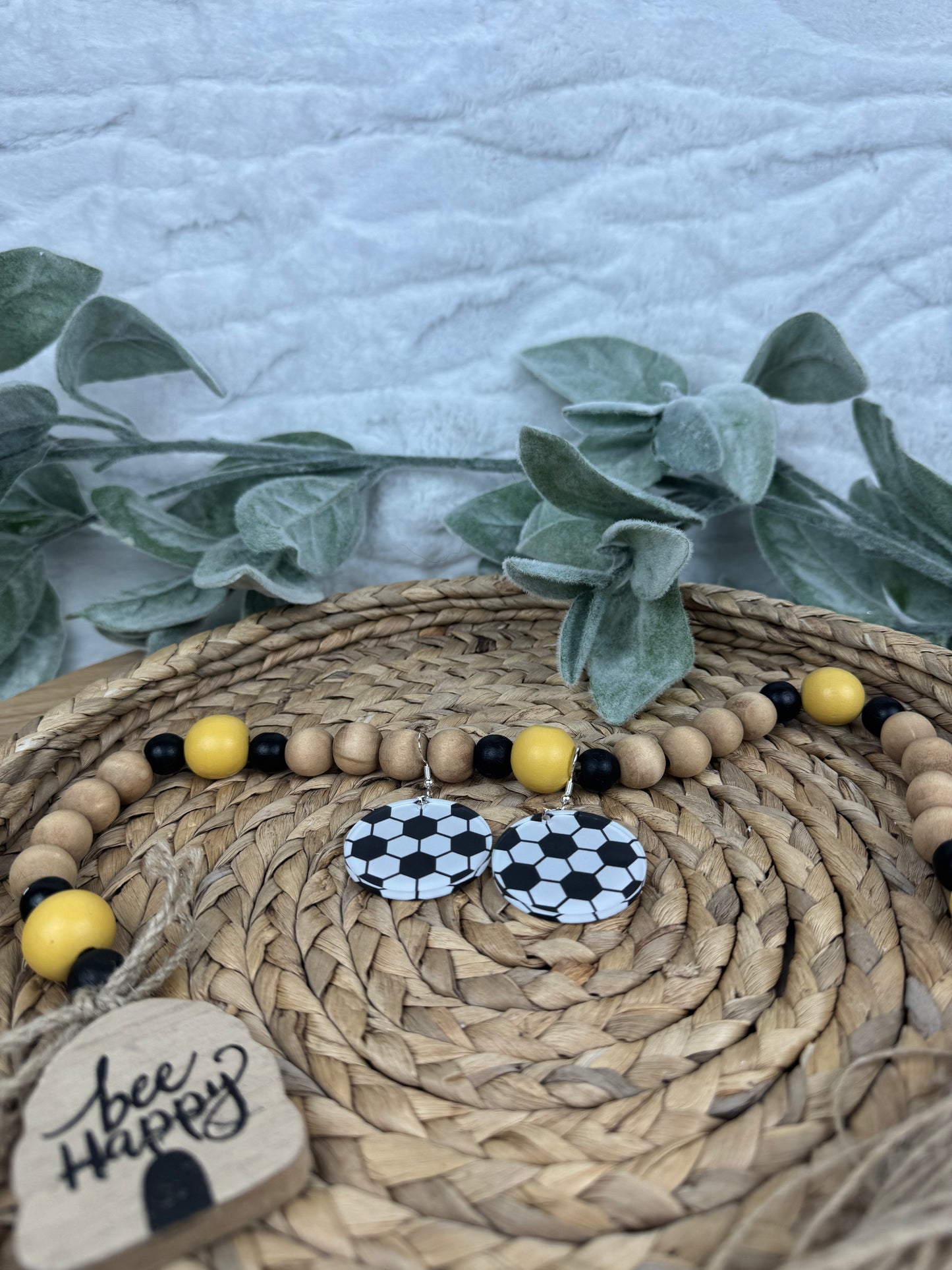 Soccer-themed dangle earrings with a black and white design, surrounded by greenery and decorative beads on a woven basket.