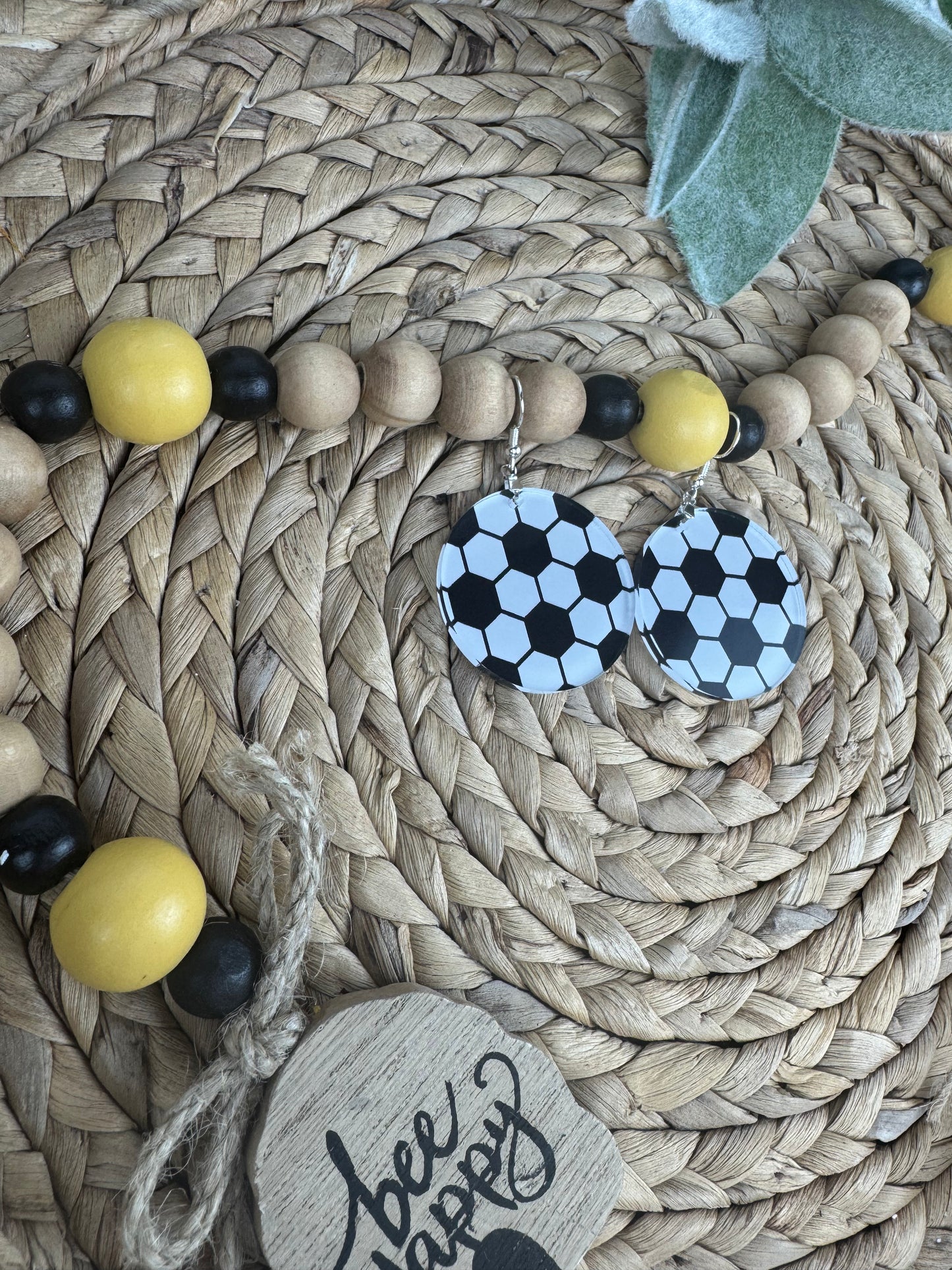 Close-up of black and white soccer ball earrings placed on a woven basket with decorative wooden beads, highlighting their sporty charm.