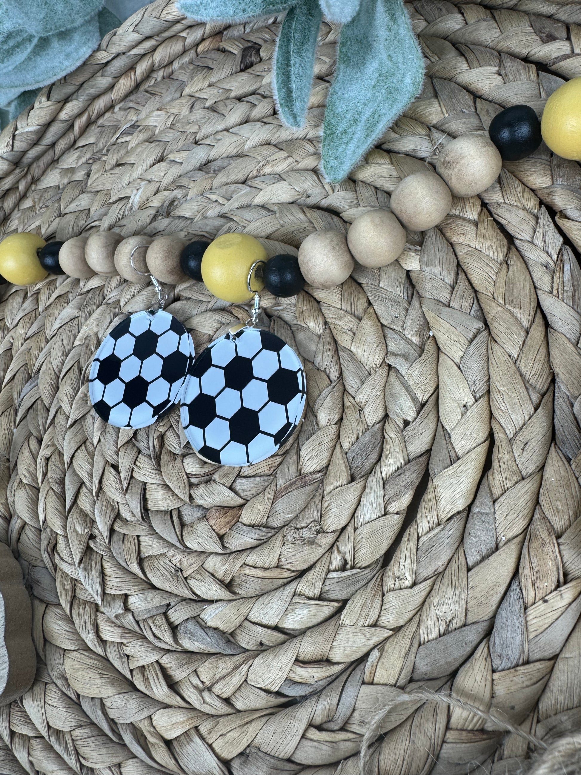 Soccer-inspired dangle earrings with black and white patterns, resting on a woven basket with yellow and black bead accents.